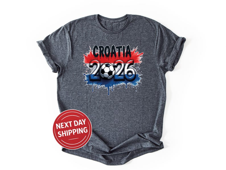 May include: A dark gray t-shirt featuring the text "CROATIA 2026" with a soccer ball graphic. The design incorporates red, white, and blue colors, with a dripping paint effect. The shirt has rolled-up sleeves.