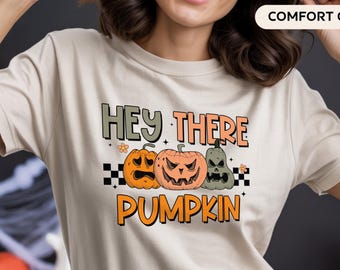 May include: Beige t-shirt with the text "HEY THERE PUMPKIN" in orange and green. The design features three cartoon pumpkins with different expressions. The shirt has a crew neck and short sleeves.