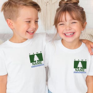 May include: Two children wearing white t-shirts with a green and white tree graphic. The shirts have the word "CAMP" above the tree and "NAME" below, with a blue wavy line. The children are smiling.