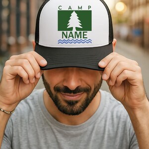 May include: A black and white trucker hat with a white front panel. The panel displays the word "CAMP" above a green rectangle containing a tree graphic and the word "NAME" below. A blue wavy line is beneath "NAME".