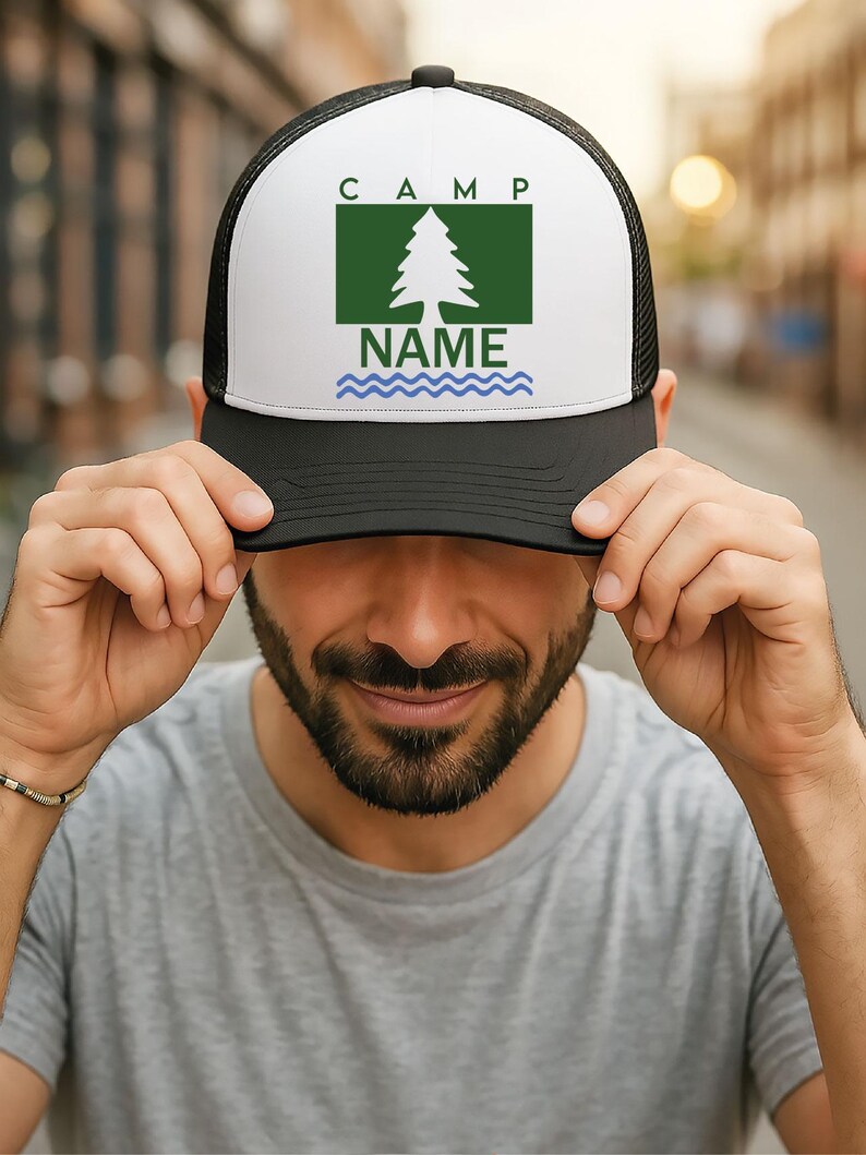 May include: A black and white trucker hat with a white front panel. The panel displays the word "CAMP" above a green rectangle containing a tree graphic and the word "NAME" below. A blue wavy line is beneath "NAME".