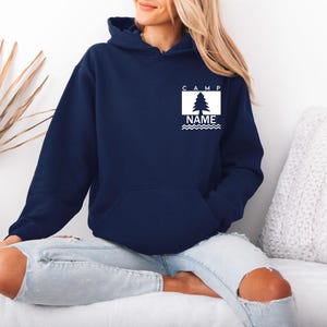 Puede incluir: Una sudadera con capucha azul marino con un bolsillo delantero. La sudadera tiene un gr&aacute;fico rectangular blanco con un &aacute;rbol y la palabra "CAMP" encima, y "NAME" debajo. El gr&aacute;fico tiene un dise&ntilde;o de l&iacute;nea ondulada.