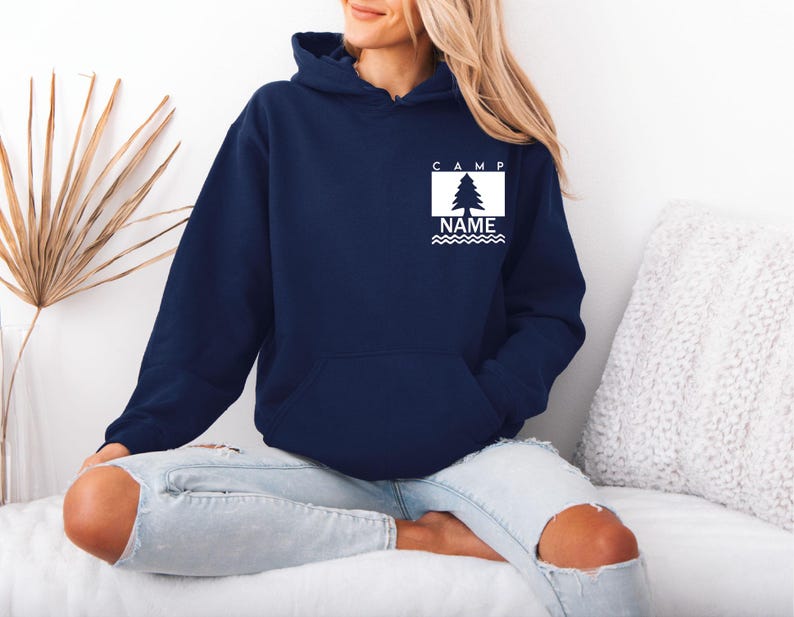 Puede incluir: Una sudadera con capucha azul marino con un bolsillo delantero. La sudadera tiene un gr&aacute;fico rectangular blanco con un &aacute;rbol y la palabra "CAMP" encima, y "NAME" debajo. El gr&aacute;fico tiene un dise&ntilde;o de l&iacute;nea ondulada.