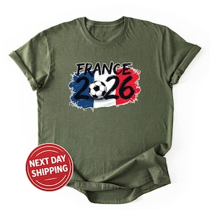 K&ouml;nnte beinhalten: Olivgr&uuml;nes T-Shirt mit einem Grafikdesign. Das Design enth&auml;lt den Text "FRANCE 2026" in Schwarz, einen Fu&szlig;ball und einen Hintergrund in den Farben der franz&ouml;sischen Flagge. Ein roter Kreis mit "NEXT DAY SHIPPING" ist ebenfalls sichtbar.