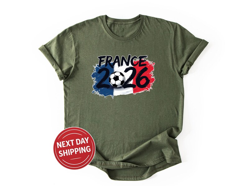 K&ouml;nnte beinhalten: Olivgr&uuml;nes T-Shirt mit einem Grafikdesign. Das Design enth&auml;lt den Text "FRANCE 2026" in Schwarz, einen Fu&szlig;ball und einen Hintergrund in den Farben der franz&ouml;sischen Flagge. Ein roter Kreis mit "NEXT DAY SHIPPING" ist ebenfalls sichtbar.