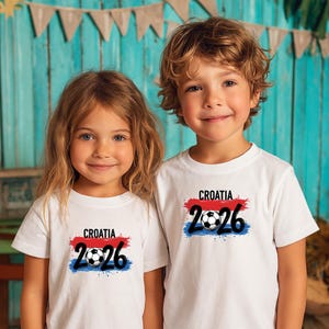 以下が含まれることがあります： 白いTシャツを着た2人の子供たち。各シャツには「CROATIA」の文字と数字「2026」、サッカーボールのグラフィックが赤と青のペイントストロークの上に配置されています。子供たちは笑顔です。