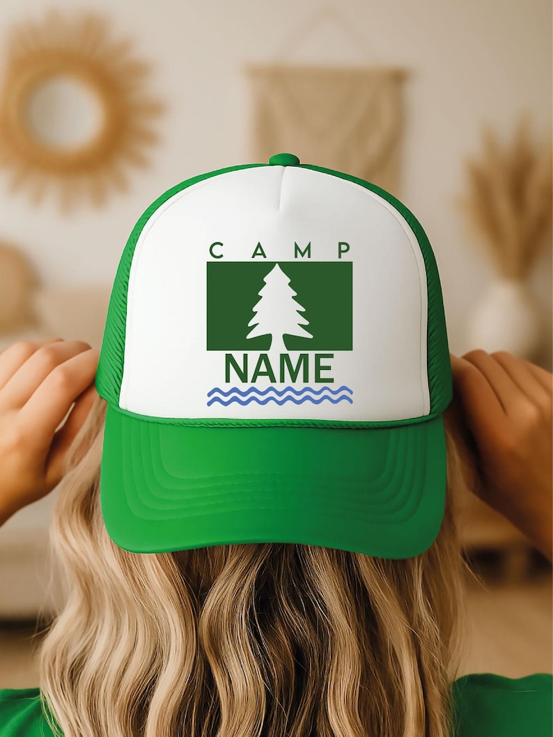 May include: A green and white trucker hat with the word "CAMP" above a green rectangle with a tree graphic. Below the tree is the word "NAME" above blue wavy lines. The hat is being held by a person.