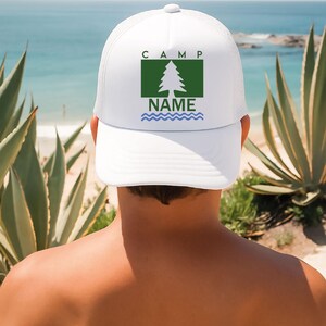 May include: White trucker hat featuring a green square with a tree graphic, the word "CAMP" above and "NAME" below. Blue wavy lines are at the bottom of the graphic. The hat is worn by a person with a beach background.