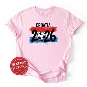 以下が含まれることがあります： 「CROATIA」の文字とサッカーボール、数字の「2026」がプリントされた薄いピンク色のTシャツ。デザインには赤、白、青の筆致が用いられ、サッカーボールは白と黒です。