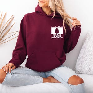 Puede incluir: Una sudadera con capucha de color granate con un bolsillo delantero y un gr&aacute;fico blanco en el pecho. El gr&aacute;fico incluye la palabra "CAMP", un &aacute;rbol, la palabra "NAME" y l&iacute;neas onduladas. La sudadera con capucha la lleva una persona.