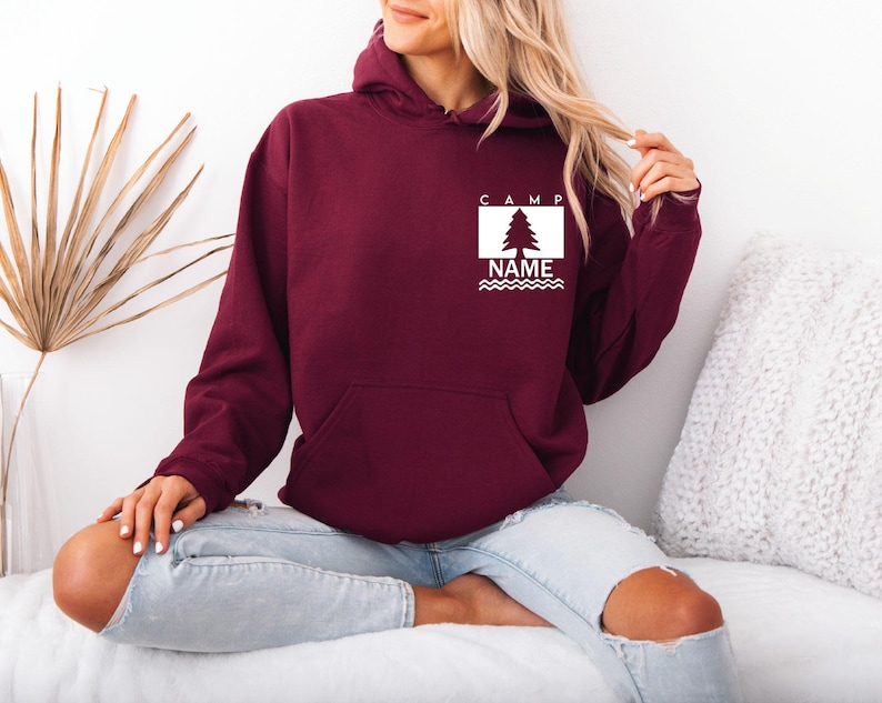 Puede incluir: Una sudadera con capucha de color granate con un bolsillo delantero y un gr&aacute;fico blanco en el pecho. El gr&aacute;fico incluye la palabra "CAMP", un &aacute;rbol, la palabra "NAME" y l&iacute;neas onduladas. La sudadera con capucha la lleva una persona.