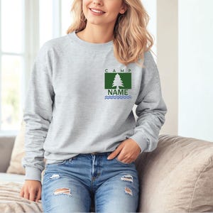 Puede incluir: Una sudadera gris claro con un gr&aacute;fico de &aacute;rbol verde y blanco y el texto "CAMP NAME". La sudadera est&aacute; hecha de un material suave y tiene mangas largas. El gr&aacute;fico incluye un &aacute;rbol, la palabra "CAMP" y la palabra "NAME".