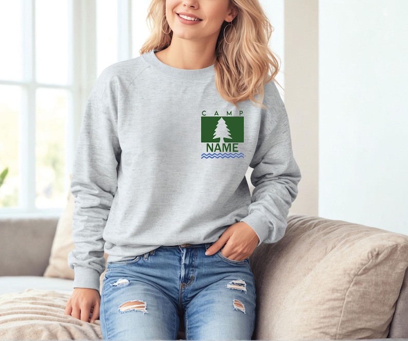Puede incluir: Una sudadera gris claro con un gr&aacute;fico de &aacute;rbol verde y blanco y el texto "CAMP NAME". La sudadera est&aacute; hecha de un material suave y tiene mangas largas. El gr&aacute;fico incluye un &aacute;rbol, la palabra "CAMP" y la palabra "NAME".