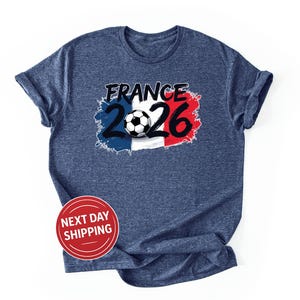 K&ouml;nnte beinhalten: Ein marineblaues T-Shirt mit einer Grafik mit dem Wort "FRANCE" in Schwarz, den Zahlen "2026" in Blau und Rot und einem Fu&szlig;ball. Das Design ist vor dem Hintergrund der franz&ouml;sischen Flagge platziert. Ein roter Kreis mit den Worten "NEXT DAY SHIPPING" befindet sich unten.