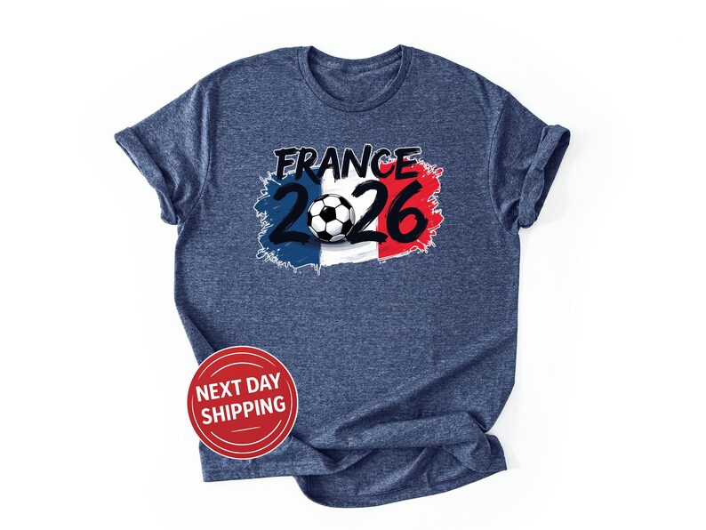 K&ouml;nnte beinhalten: Ein marineblaues T-Shirt mit einer Grafik mit dem Wort "FRANCE" in Schwarz, den Zahlen "2026" in Blau und Rot und einem Fu&szlig;ball. Das Design ist vor dem Hintergrund der franz&ouml;sischen Flagge platziert. Ein roter Kreis mit den Worten "NEXT DAY SHIPPING" befindet sich unten.