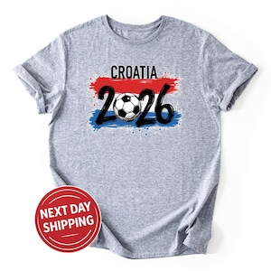 以下が含まれることがあります： 「CROATIA」の文字と「2026」の数字、サッカーボールのグラフィックがプリントされたヘザーグレーのTシャツ。赤、白、青の筆致のデザインは、国旗を連想させます。カジュアルウェアです。