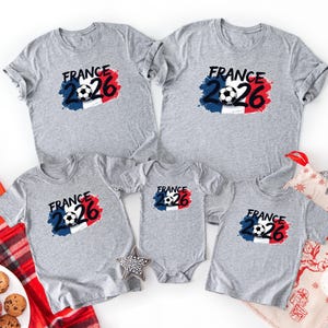 K&ouml;nnte beinhalten: Eine Auswahl grauer T-Shirts und ein Baby-Body mit einem Fu&szlig;ball-Design und dem Text "FRANCE 26" in Schwarz, Rot und Blau. Die Shirts sind kurz&auml;rmlig und eignen sich f&uuml;r eine Familie oder Gruppe.