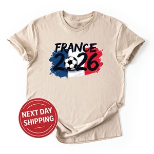 K&ouml;nnte beinhalten: Beiges T-Shirt mit einem Grafikdesign. Das Design beinhaltet das Wort "France" in Schwarz, die Zahl "2026" und einen Fu&szlig;ball, vor einem blau-wei&szlig;-roten Hintergrund. Das T-Shirt hat kurze &Auml;rmel und einen Rundhalsausschnitt.