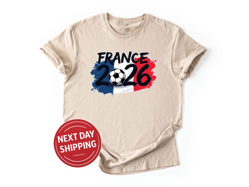 K&ouml;nnte beinhalten: Beiges T-Shirt mit einem Grafikdesign. Das Design beinhaltet das Wort "France" in Schwarz, die Zahl "2026" und einen Fu&szlig;ball, vor einem blau-wei&szlig;-roten Hintergrund. Das T-Shirt hat kurze &Auml;rmel und einen Rundhalsausschnitt.