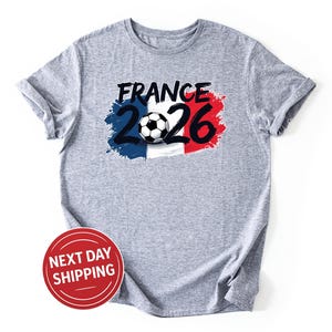 K&ouml;nnte beinhalten: Ein meliertes graues T-Shirt mit dem Schriftzug "FRANCE 2026" in Schwarz und Wei&szlig;, mit einem Fu&szlig;ball-Motiv. Das Design integriert die Farben der franz&ouml;sischen Flagge: Blau, Wei&szlig; und Rot. Das T-Shirt hat hochgekrempelte &Auml;rmel.
