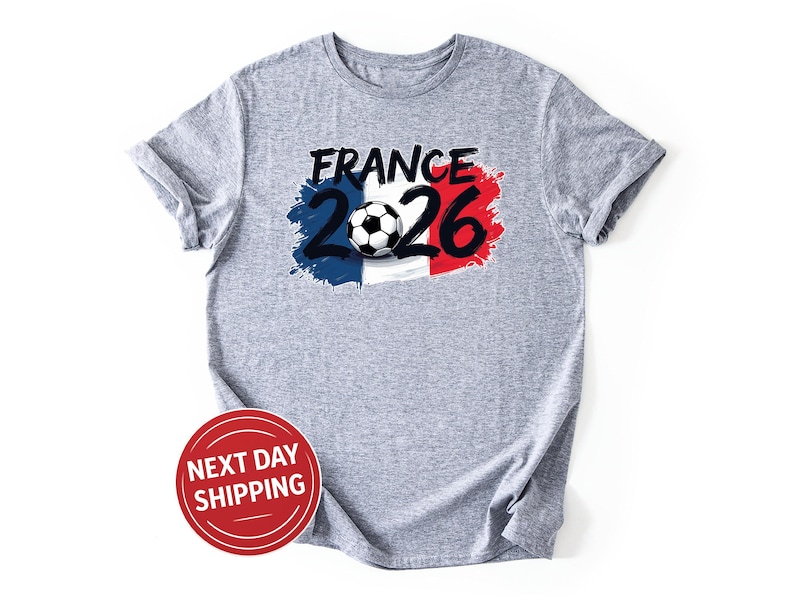 K&ouml;nnte beinhalten: Ein meliertes graues T-Shirt mit dem Schriftzug "FRANCE 2026" in Schwarz und Wei&szlig;, mit einem Fu&szlig;ball-Motiv. Das Design integriert die Farben der franz&ouml;sischen Flagge: Blau, Wei&szlig; und Rot. Das T-Shirt hat hochgekrempelte &Auml;rmel.