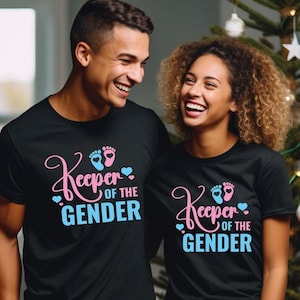 May include: Two people wearing black t-shirts with the text "Keeper of the Gender" and a pink and blue footprint graphic. The couple is smiling and looking at each other.