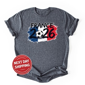 K&ouml;nnte beinhalten: Dunkelgraues T-Shirt mit einem Aufdruck, der das Wort "FRANCE" &uuml;ber der Zahl "2026" und einen Fu&szlig;ball zeigt, vor dem Hintergrund der franz&ouml;sischen Flaggenfarben. Das T-Shirt hat kurze &Auml;rmel.