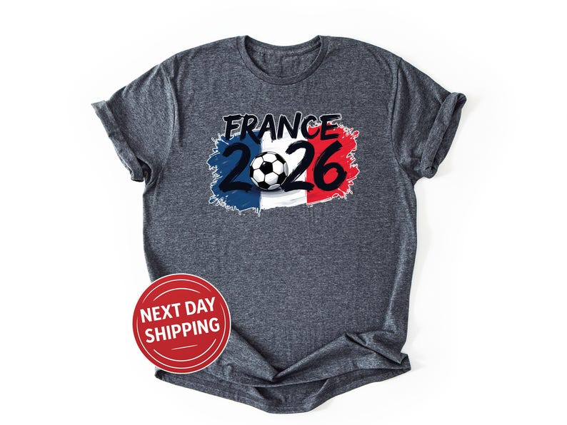 K&ouml;nnte beinhalten: Dunkelgraues T-Shirt mit einem Aufdruck, der das Wort "FRANCE" &uuml;ber der Zahl "2026" und einen Fu&szlig;ball zeigt, vor dem Hintergrund der franz&ouml;sischen Flaggenfarben. Das T-Shirt hat kurze &Auml;rmel.