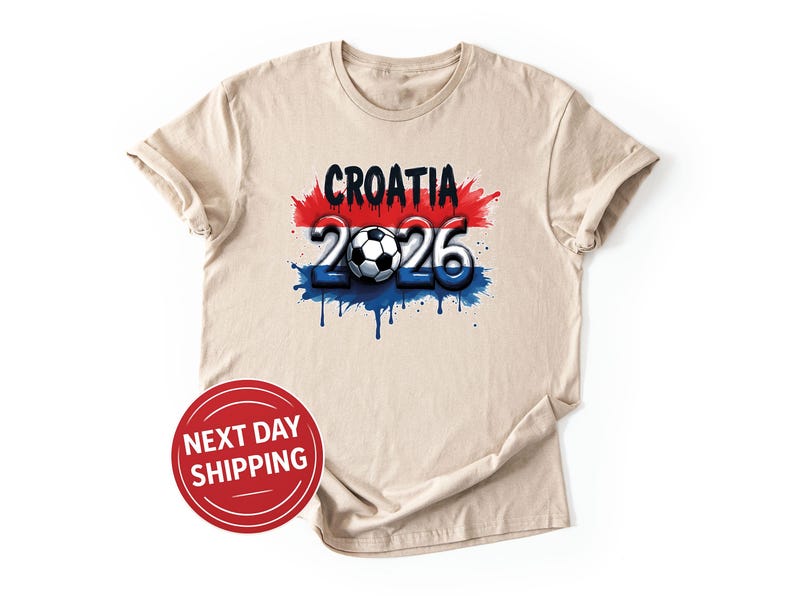 May include: A beige t-shirt featuring the word "CROATIA" in black above the numbers "2026" with a soccer ball in the center. The design incorporates red, white, and blue paint splatters, resembling a flag. A red circle with "NEXT DAY SHIPPING" is in the lower left.