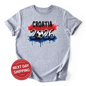 May include: A heather gray t-shirt featuring the text "CROATIA 2026" in a design with red, white, and blue accents, and a soccer ball. The design has a dripping paint effect. The shirt is a casual wear item.