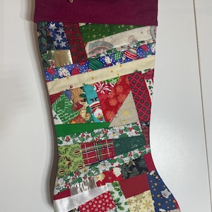 May include: A Christmas stocking featuring a patchwork design of various holiday-themed fabrics in red, green, blue, and gold. The top cuff is a solid deep maroon fabric with a gold hanging loop. The body consists of assorted geometric fabric scraps.