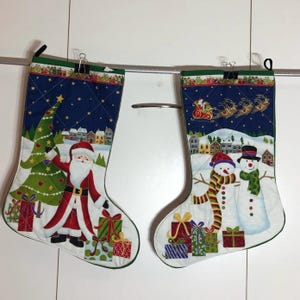 Peut inclure: Deux chaussettes de Noël en tissu matelassé. Une chaussette représente le Père Noël avec un sapin de Noël et des cadeaux. L'autre chaussette représente deux bonhommes de neige avec des cadeaux. Les deux chaussettes ont un fond bleu avec des flocons de neige blancs.