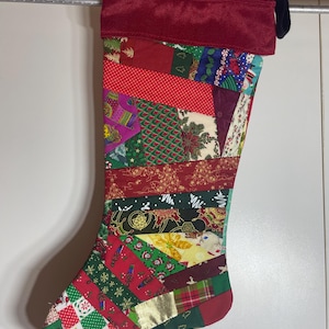 May include: A Christmas stocking featuring a patchwork design of various holiday-themed fabrics in red, green, white, and gold. The stocking has a solid dark red cuff at the top and a black hanging loop, constructed from multiple angled fabric strips.