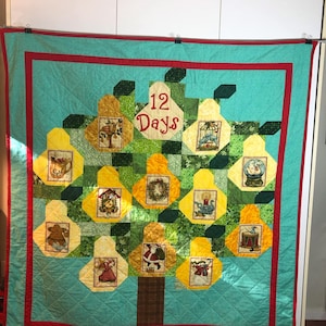 May include: A turquoise blue quilted Christmas tree with a red border. The tree is decorated with 12 yellow and green squares, each with a different Christmas scene. The tree is topped with a white tag that says "12 Days".