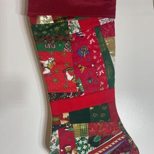 Peut inclure: Une chaussette de Noël avec un motif patchwork composé de divers tissus sur le thème des fêtes en rouge, vert et doré. Les motifs incluent des chevaux à bascule, des ours en peluche, des cadeaux et du houx. Elle possède un revers rouge uni.