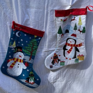 May include: Two Christmas stockings with a winter theme. One stocking features a snowman with a red scarf and a penguin on a sledge. The other stocking features a penguin with a red scarf and a snowman in a snow globe. Both stockings have a red cuff.