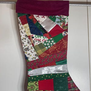 May include: A Christmas stocking featuring a patchwork design of various holiday-themed fabrics in red, green, white, and silver. The stocking has a solid dark red cuff at the top and a gold hanging loop. The fabric patterns include holly, reindeer, and the text HO HO HO.