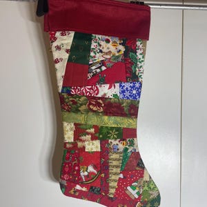 Peut inclure: Une chaussette de Noël avec un motif patchwork composé de divers tissus sur le thème des fêtes en rouge, vert, blanc et or. La chaussette présente un revers rouge foncé uni sur le haut et est faite de multiples pièces de tissu géométriques.