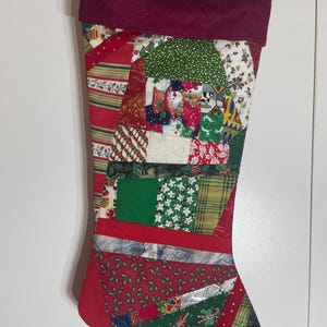 Peut inclure: Une chaussette de Noël au design patchwork composé de divers tissus sur le thème des fêtes en rouge, vert et blanc. La chaussette présente un revers rouge foncé uni sur le dessus et est faite de multiples pièces de tissu angulaires.