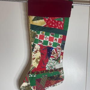 Peut inclure: Une chaussette de Noël présentant un motif en patchwork composé de divers tissus sur le thème des fêtes en rouge, vert et blanc. La chaussette possède un revers en velours rouge foncé uni sur le haut et est faite de chutes de tissu.