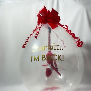 Puede incluir: Un globo de pl&aacute;stico transparente con un lazo rojo y una cinta roja. Dentro del globo hay una mu&ntilde;eca de elfo roja y blanca. El globo tiene el texto "Charlotte I'M BACK!" impreso en &eacute;l.