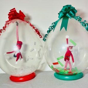 Puede incluir: Dos globos de pl&aacute;stico transparentes con lazos rojos y verdes. Cada globo contiene un peque&ntilde;o mu&ntilde;eco de elfo en su interior. Un elfo est&aacute; colgado boca abajo y el otro est&aacute; de pie con los brazos en alto.