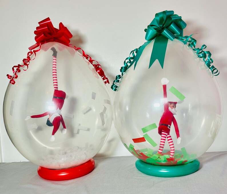 Puede incluir: Dos globos de pl&aacute;stico transparentes con lazos rojos y verdes. Cada globo contiene un peque&ntilde;o mu&ntilde;eco de elfo en su interior. Un elfo est&aacute; colgado boca abajo y el otro est&aacute; de pie con los brazos en alto.