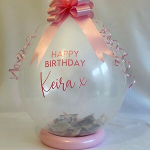 May include: A clear balloon with a pink ribbon and bow, filled with cash. The balloon has the text "HAPPY BIRTHDAY Keira x" printed on it.