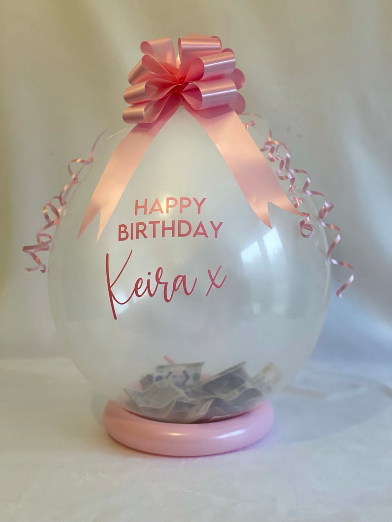 May include: A clear balloon with a pink ribbon and bow, filled with cash. The balloon has the text "HAPPY BIRTHDAY Keira x" printed on it.