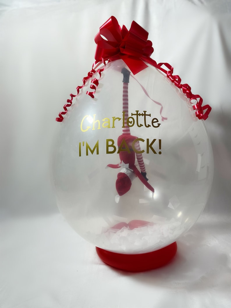 Puede incluir: Un globo de pl&aacute;stico transparente con un lazo rojo y una cinta roja. Dentro del globo hay una mu&ntilde;eca de elfo roja y blanca. El globo tiene el texto "Charlotte I'M BACK!" impreso en &eacute;l.