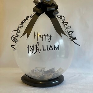 May include: A clear balloon with a black ribbon and bow, filled with cash. The balloon has the text "Happy 18th LIAM" printed on it.