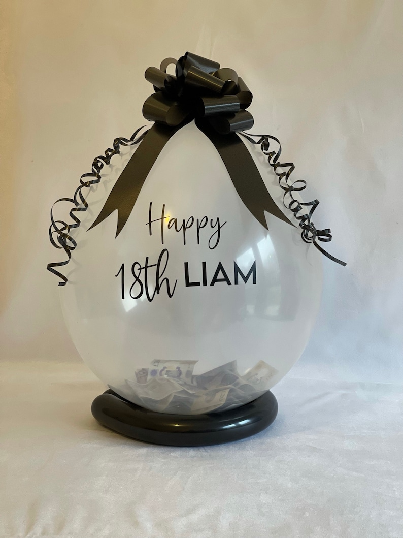 May include: A clear balloon with a black ribbon and bow, filled with cash. The balloon has the text "Happy 18th LIAM" printed on it.