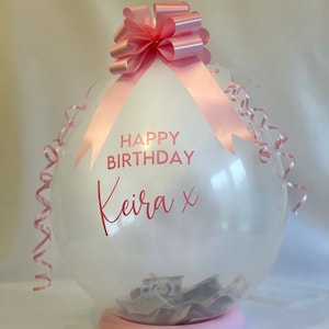 May include: A clear balloon with a pink ribbon and bow, filled with cash. The balloon has the text "HAPPY BIRTHDAY Keira x" printed on it.