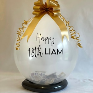 May include: A clear balloon with a gold ribbon and bow, filled with cash. The balloon has the text "Happy 18th LIAM" printed on it.